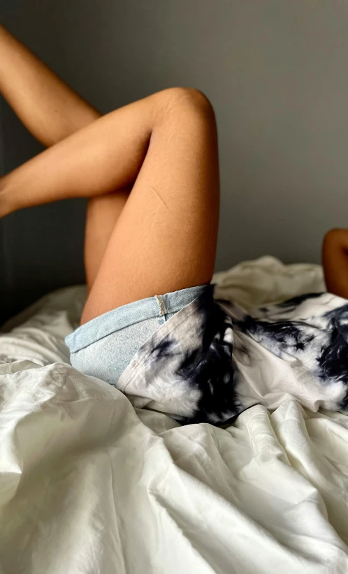 a woman laying on a bed with her legs up, by Robbie Trevino, happening, wearing denim short shorts, wearing a tie-dye t-shirt, close up shot from the top, high details photo