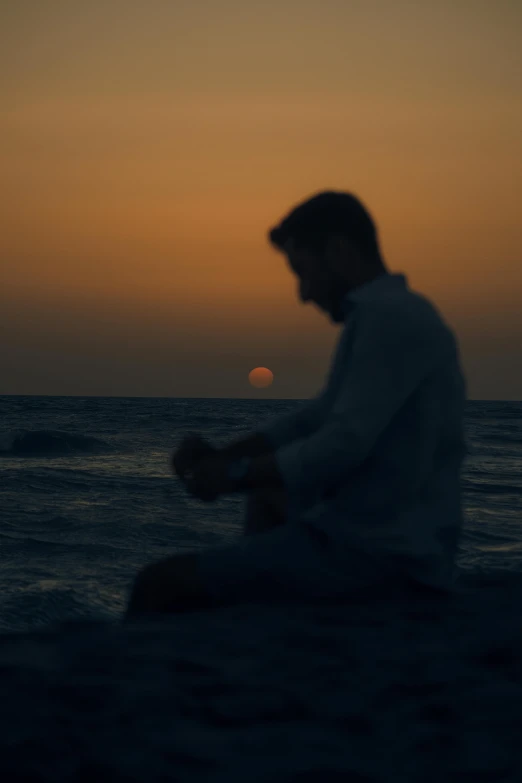 a man sitting on top of a beach next to the ocean, a picture, pexels contest winner, romanticism, praying at the sun, profile image, low light cinematic, still from a music video