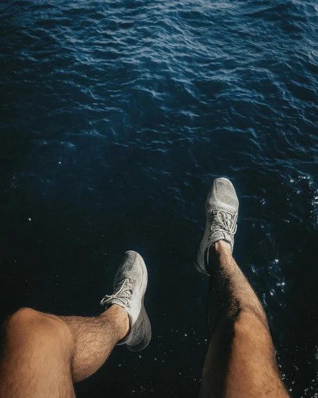 a man sitting on top of a boat next to a body of water, by Adam Dario Keel, pexels contest winner, gray shorts and black socks, two male, legs taking your pov, wearing white sneakers