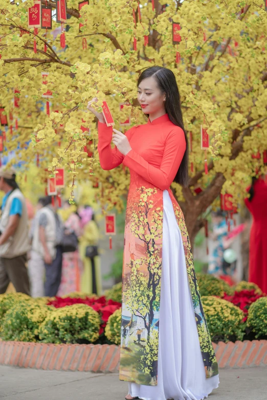 a woman standing in front of a tree holding a cell phone, inspired by Cui Bai, pexels contest winner, ao dai, festive colors, square, panoramic view of girl