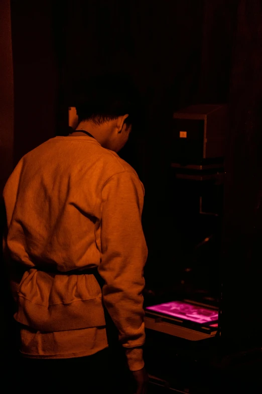 a man standing in front of a tv in a dark room, a silk screen, unsplash, holography, wearing a pink hoodie, capsule hotel, digging, brown atmospheric lighting