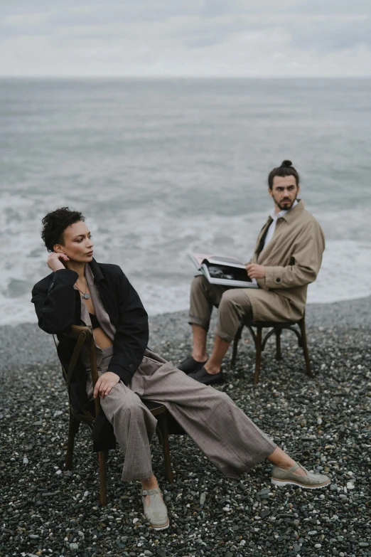 a man and a woman sitting next to each other on a beach, unsplash, renaissance, trench coat and suit, ekaterina, overcast, black sand