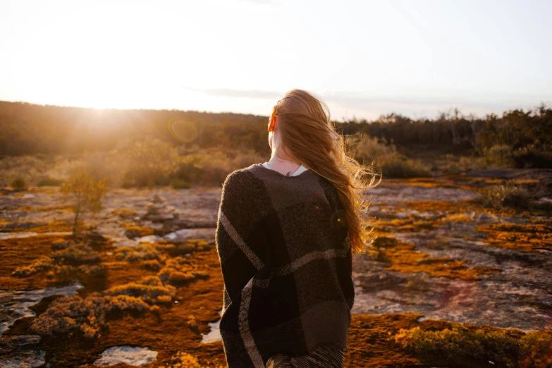 a woman standing on top of a rocky hill, by Lee Loughridge, pexels contest winner, flowing backlit hair, in the australian outback, looking from behind, wearing desert poncho