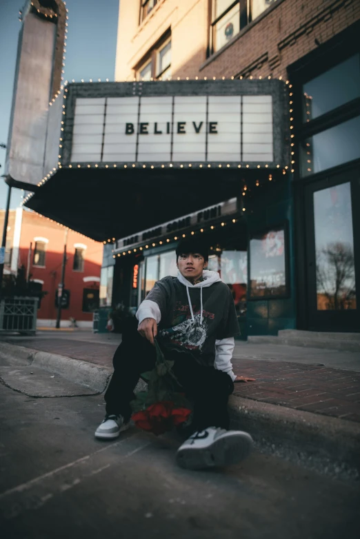 a man sitting on a skateboard in front of a movie theater, an album cover, unsplash contest winner, wearing a hoodie and flowers, confident pose, he can ’ t believe his eyes, bella poarch