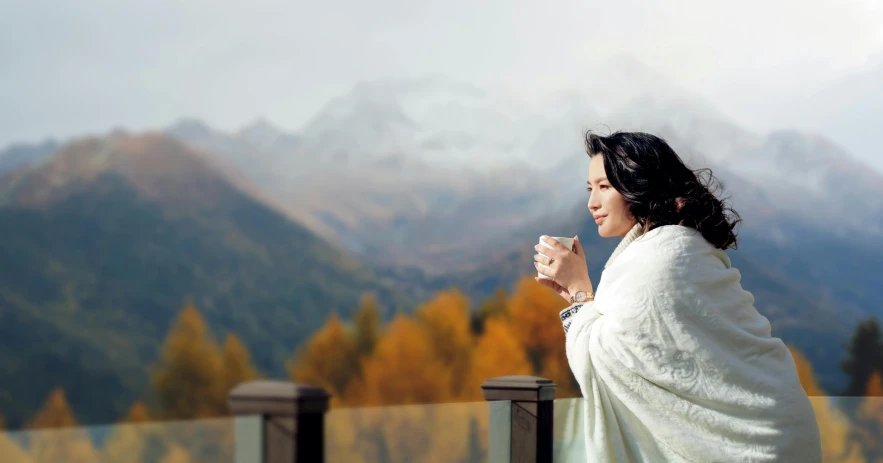 a woman wrapped in a blanket drinking a cup of coffee, inspired by Zhou Wenjing, pexels contest winner, mountains on the background, autumnal empress, marina abramovic, at the terrace