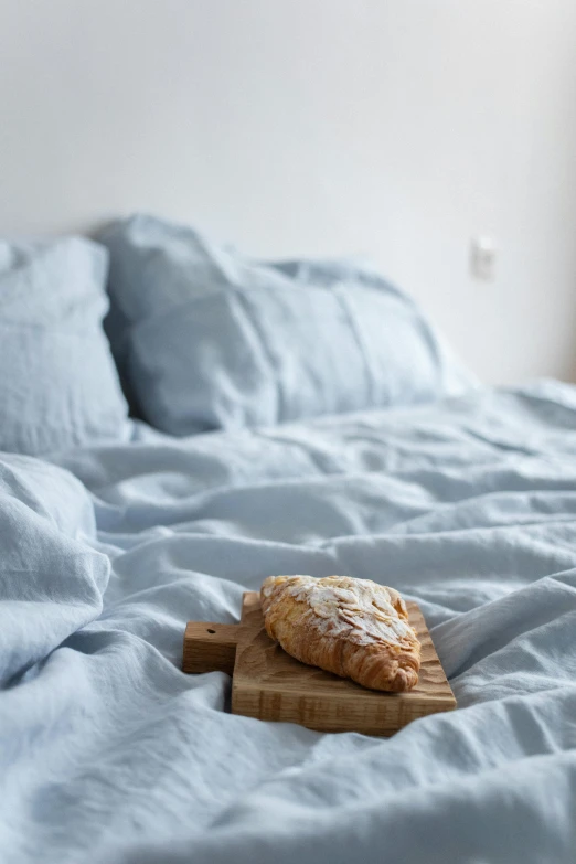 a wooden cutting board sitting on top of a bed, by Simon Gaon, pastel blue, bakery, breakfast, linen