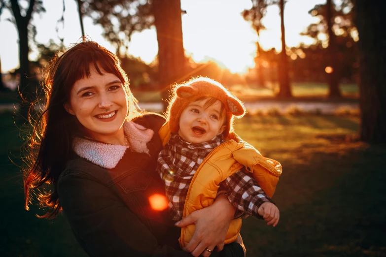 a woman holding a baby in her arms, pexels contest winner, golden hour 8k, happily smiling at the camera, avatar image, sunny day in a park