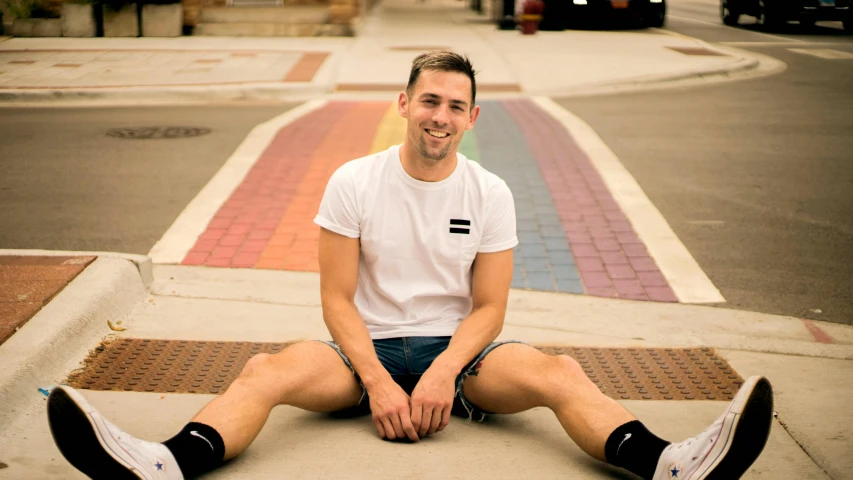 a man sitting on the curb of a city street, inspired by Ryan Pancoast, happening, pride month, wearing shorts and t shirt, lachlan bailey, headshot photo