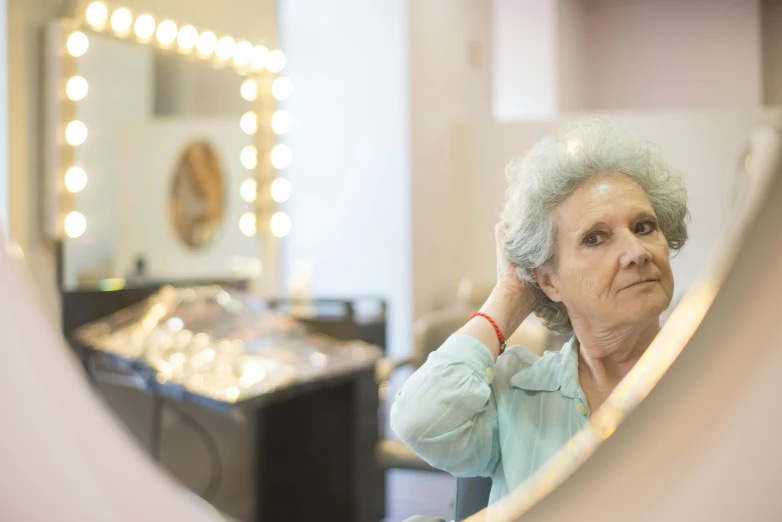 a woman combing her hair in front of a mirror, by Pamela Drew, pexels contest winner, elderly woman, theater dressing room, glowing with silver light, looking to his left