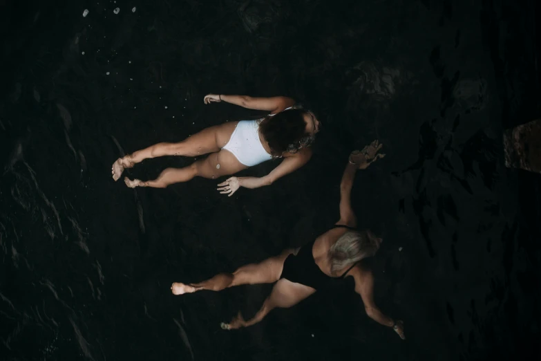 a couple of people laying on top of a body of water, by Emma Andijewska, unsplash contest winner, minimalism, on a dark rock background, is wearing a swimsuit, twins, high angle view