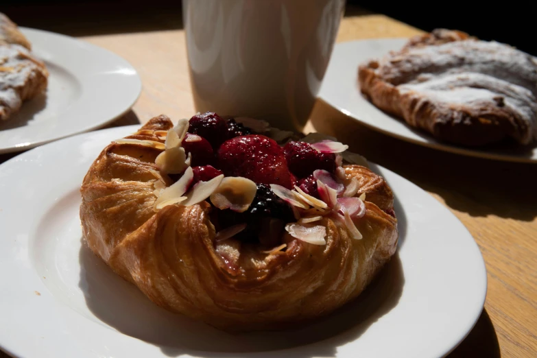 a pastry sitting on top of a white plate next to a cup of coffee, inspired by Richmond Barthé, unsplash, renaissance, plates of fruit, a wooden, sparkling in the sunlight, flowers around