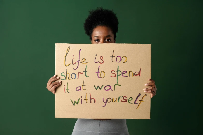 a woman holding a sign that says life is too short to spend it's all war with yourself, a picture, trending on pexels, sots art, dark-skinned, cut out of cardboard, promotional image, profile picture
