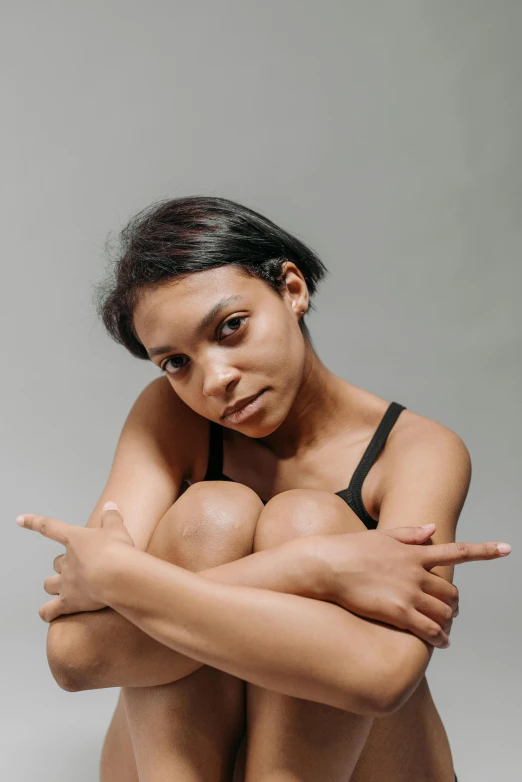 a woman sitting on the ground with her legs crossed, antipodeans, depressed dramatic bicep pose, with brown skin, pointing at the camera, on a pale background