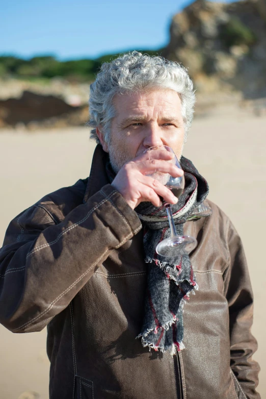 a man standing on top of a sandy beach, holding a glass of wine, dark grey haired man, mouldy juice, actor