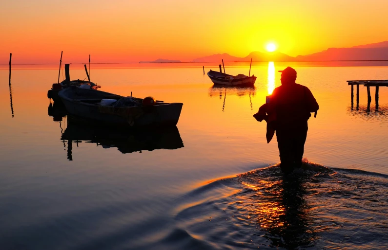 a man standing in the middle of a body of water, fishing boats, vibrant sunrise, photograph of the year, slide show