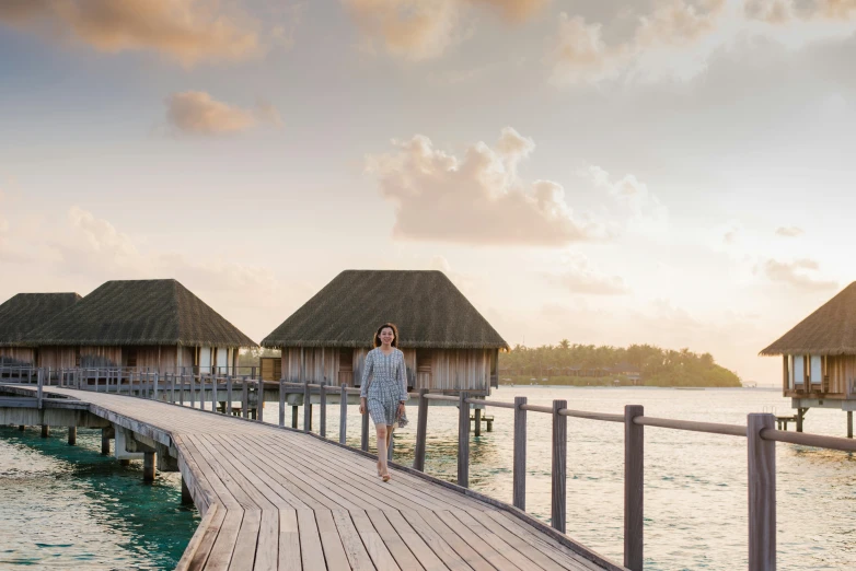 a man walking on a boardwalk over a body of water, maldives in background, huts, golden hour time, 40 years old women