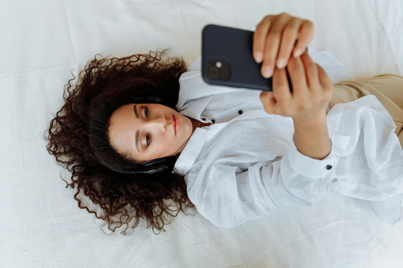 a woman laying on a bed taking a picture with her cell phone, an album cover, trending on pexels, brown curly hair, wearing gaming headset, wearing a white button up shirt, thumbnail