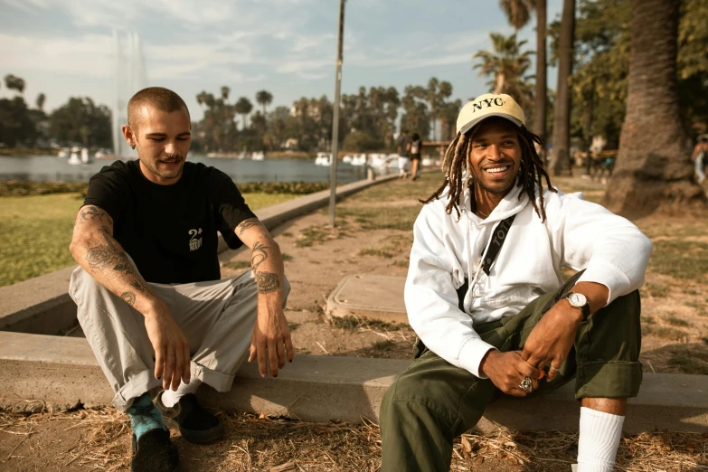 a couple of men sitting next to each other, a portrait, by Stokely Webster, unsplash, long beach background, bowater charlie and brom gerald, both smiling for the camera, wearing cargo pants