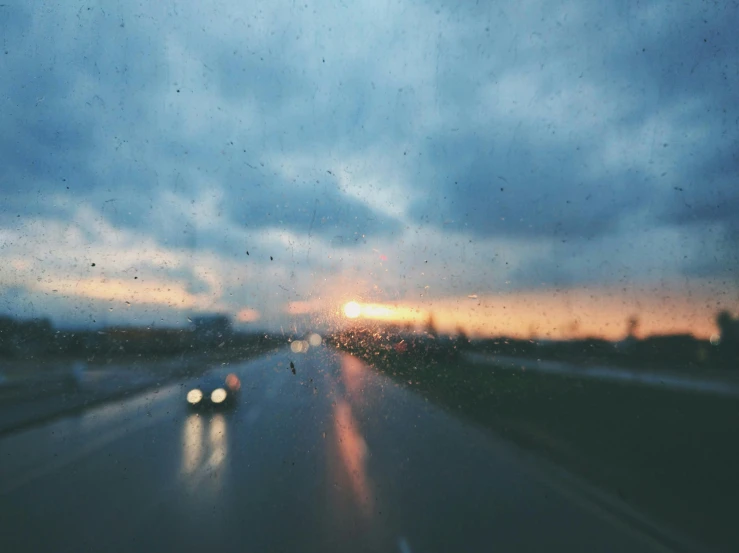 a car driving down the road on a rainy day, inspired by Elsa Bleda, pexels contest winner, visual art, sunset with cloudy skies, the sun reflecting on a window, blurred, vibrant but dreary blue