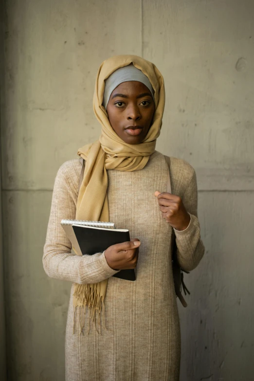 a woman standing in front of a wall holding a book, inspired by Maryam Hashemi, trending on unsplash, wearing turtleneck, dark skin tone, standing in class, head scarf