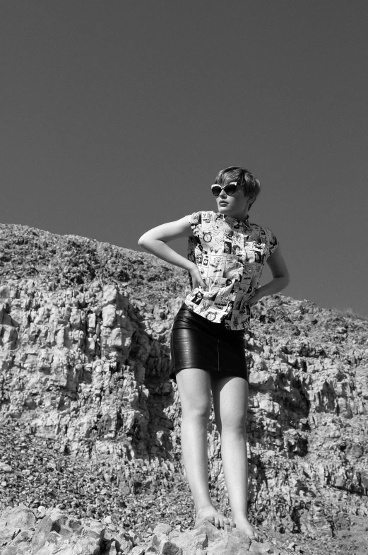 a woman standing on top of a rocky hill, a black and white photo, inspired by Cindy Sherman, with sunglass, wearing honey - themed miniskirt, 60's sci-fi pinup style, myself