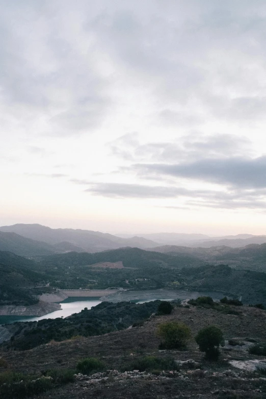 a man standing on top of a hill next to a lake, by Alexis Grimou, distant town in valley and hills, overcast dusk, overview, marbella landscape