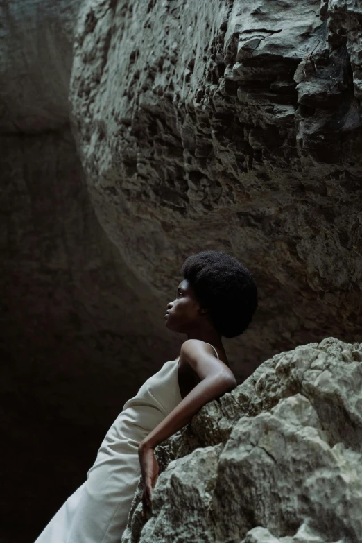 a woman in a white dress sitting on a rock, inspired by Gordon Parks, unsplash, afrofuturism, caves, natural hair, 1960s color photograph, ignant