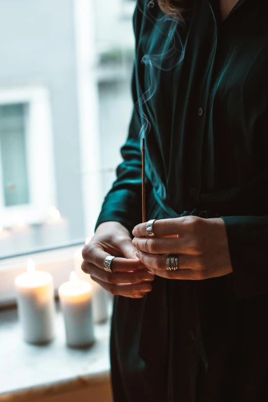 a woman standing in front of a window holding a candle, by Christen Dalsgaard, trending on unsplash, wearing two metallic rings, wearing a grey robe, smoking a joint, wearing two silver bracelets