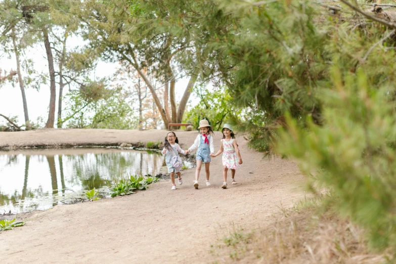 a group of children walking down a dirt road next to a body of water, walking at the garden, photoshoot for skincare brand, threes, waterlily pond