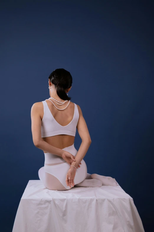 a woman sitting on top of a white table, a marble sculpture, inspired by Fei Danxu, unsplash, massurrealism, strings of pearls, marjaryasana and bitilasana, relaxed. blue background, showing her shoulder from back