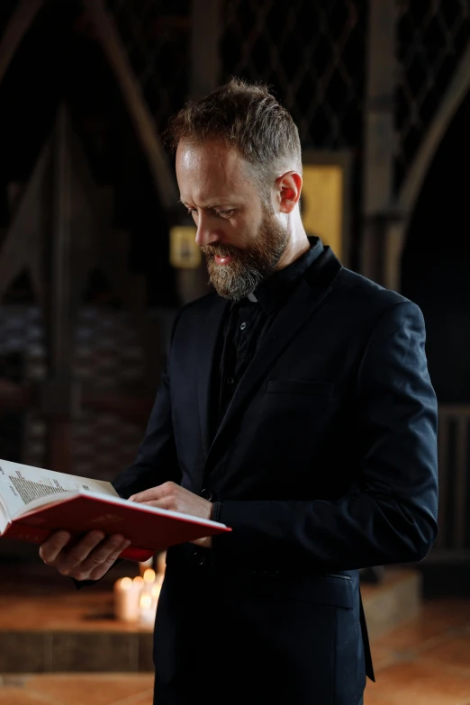 a man in a suit reading a book, inspired by Thomas Tudor, in church, with a beard and a black shirt, press shot, holy ceremony