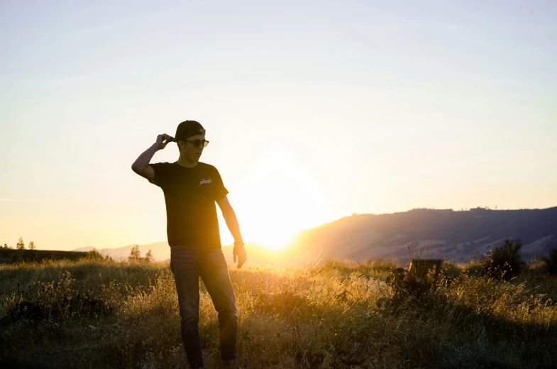 a man standing on top of a grass covered field, sunset glow around head, wearing sunglasses and a cap, carson ellis, avatar image