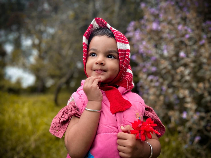 a little girl that is standing in the grass, by Almada Negreiros, pixabay contest winner, turban of vibrant flowers, wearing a red gilet, wearing a pink hoodie, 🤤 girl portrait