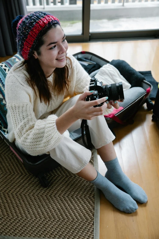 a woman sitting on the floor holding a camera, luggage, wearing kneesocks, grey, smiling slightly