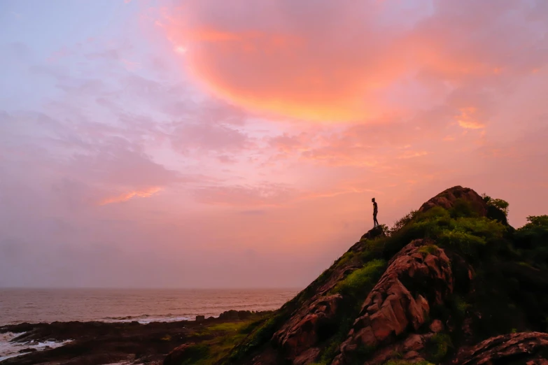 a person standing on top of a hill next to the ocean, pexels contest winner, pink clouds, red sandstone natural sculptures, indiecraft aesthetic, in the evening