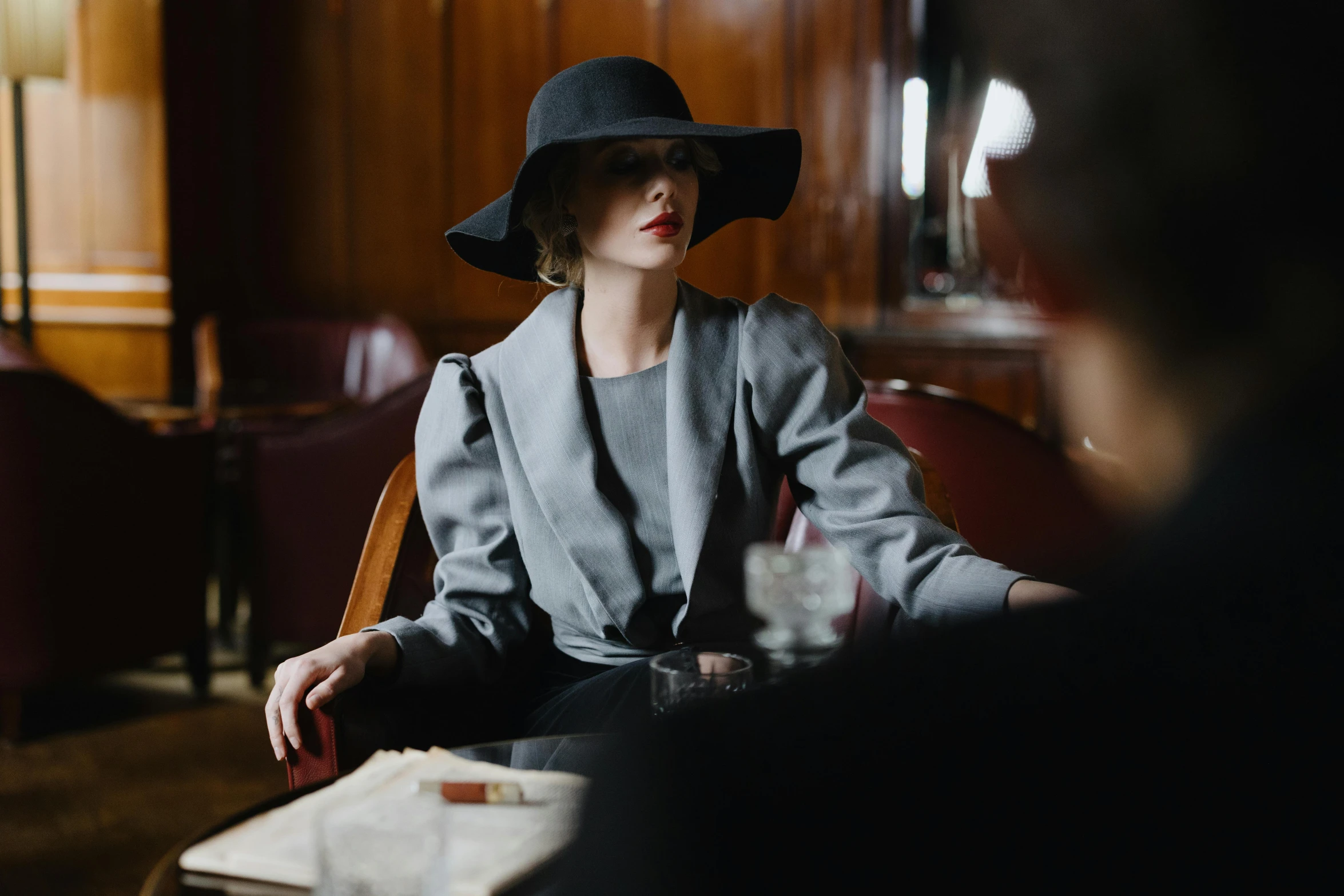 a woman sitting in a chair with a hat on, by Emma Andijewska, pexels contest winner, wearing dark grey suit, scene from a dinner party, wearing jacket and skirt, woman model