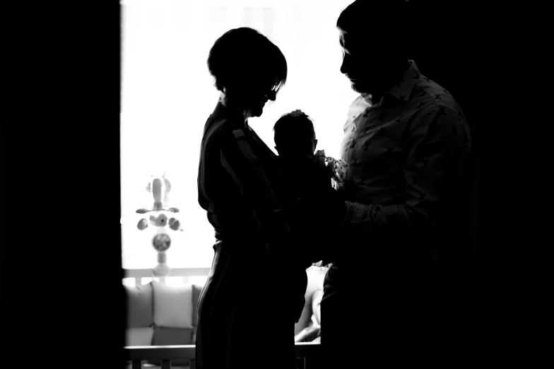 a man standing next to a woman holding a baby, a black and white photo, by Miroslava Sviridova, pexels, silhouette :7, window light, instagram post, fan favorite