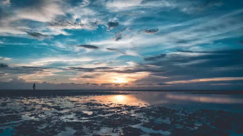 a person standing on top of a beach under a cloudy sky, it is sunset, shallow water, unsplash photography, panorama view of the sky