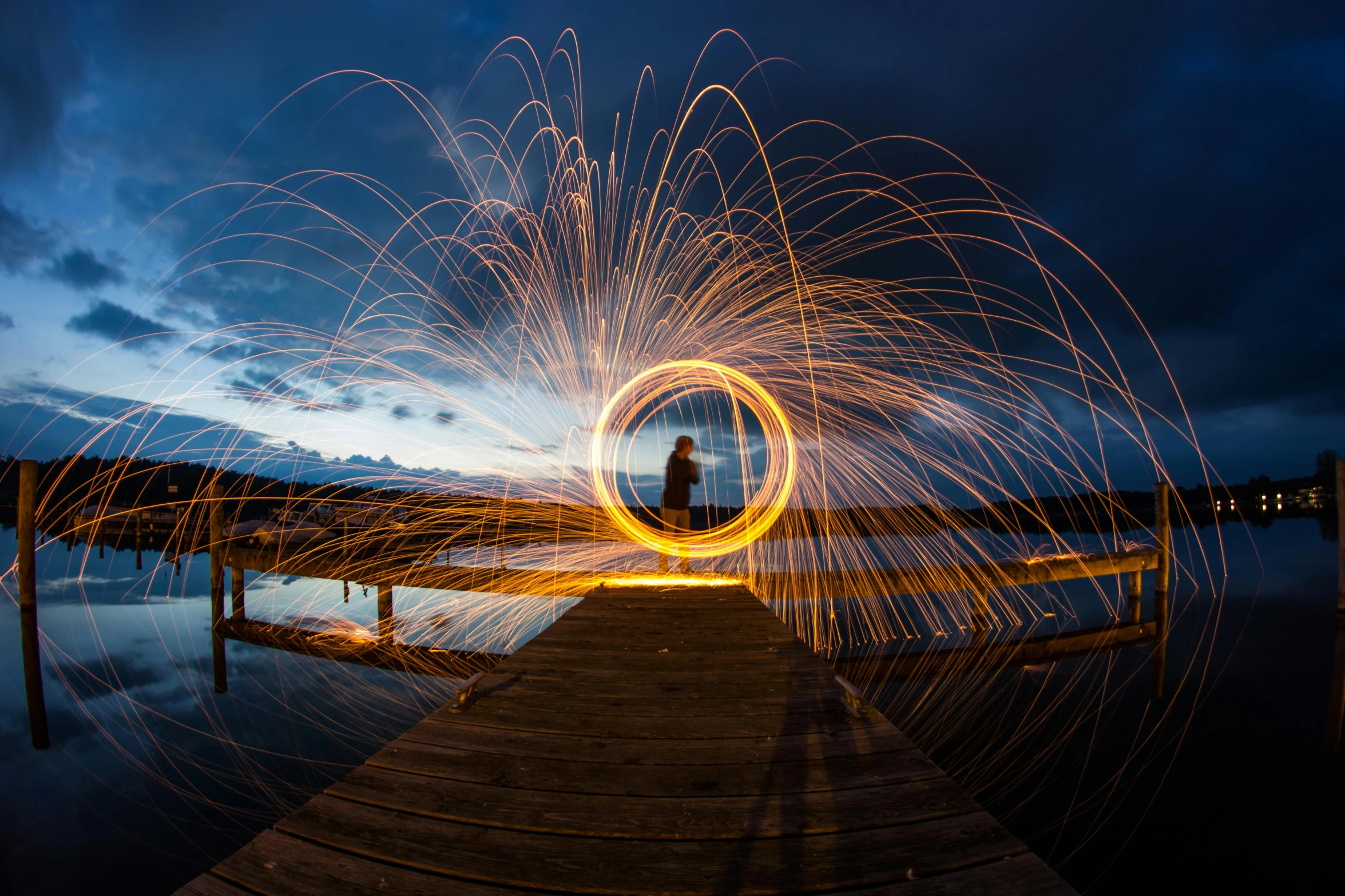 a man standing on top of a wooden pier next to a body of water, by Sebastian Spreng, pexels contest winner, sparklers, spiraling, circle, by greg rutkowski