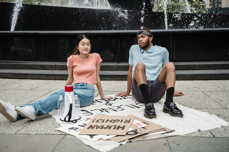 a man and woman sitting on the ground in front of a fountain, by Nina Hamnett, trending on unsplash, graffiti, protesters holding placards, outlive streetwear collection, rugs, spilled milk