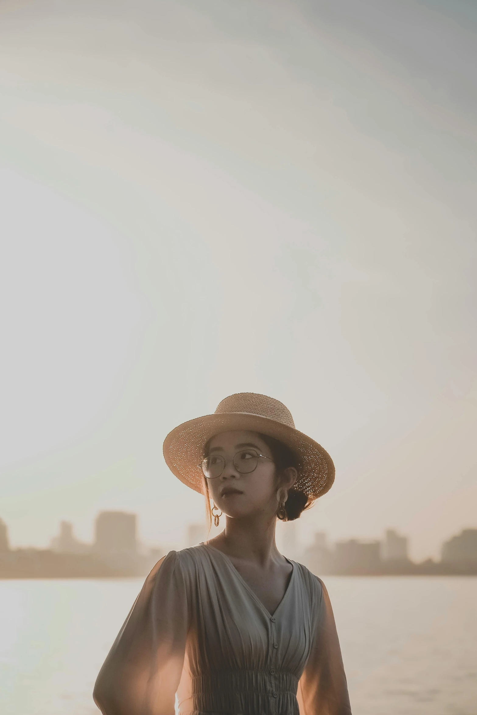 a woman standing in front of a body of water, by Lily Delissa Joseph, pexels contest winner, caracter with brown hat, portrait of modern darna, summer evening, pale beige sky