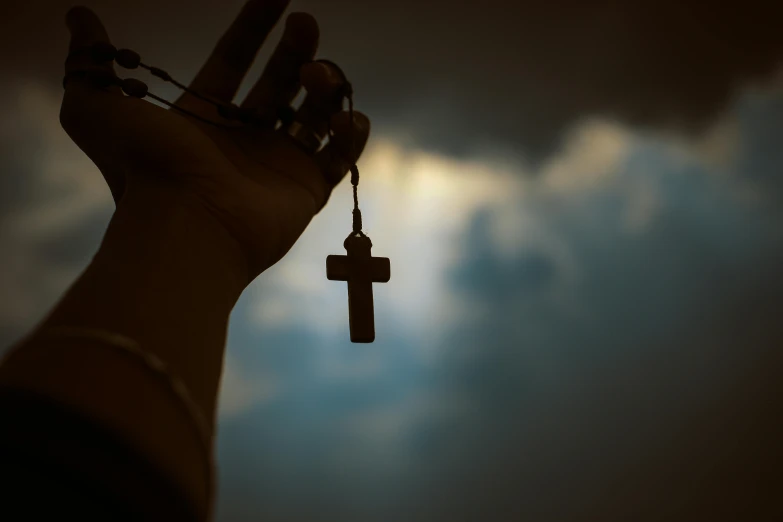 a person holding a cross in front of a cloudy sky, unsplash, hurufiyya, wooden jewerly, hanging from the ceiling, high quality product image”, silhouetted