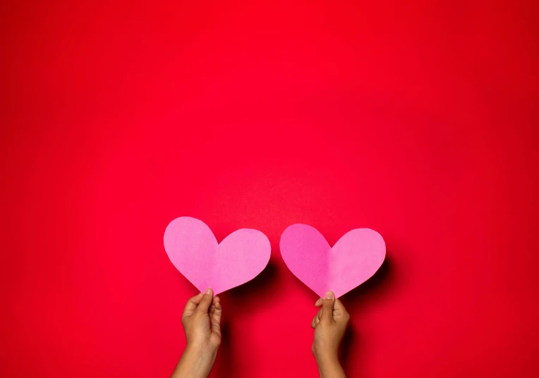 two hands holding pink paper hearts against a red background, by Julia Pishtar, pexels contest winner, minimalism, plain background, very large, holding a stuff, adult pair of twins