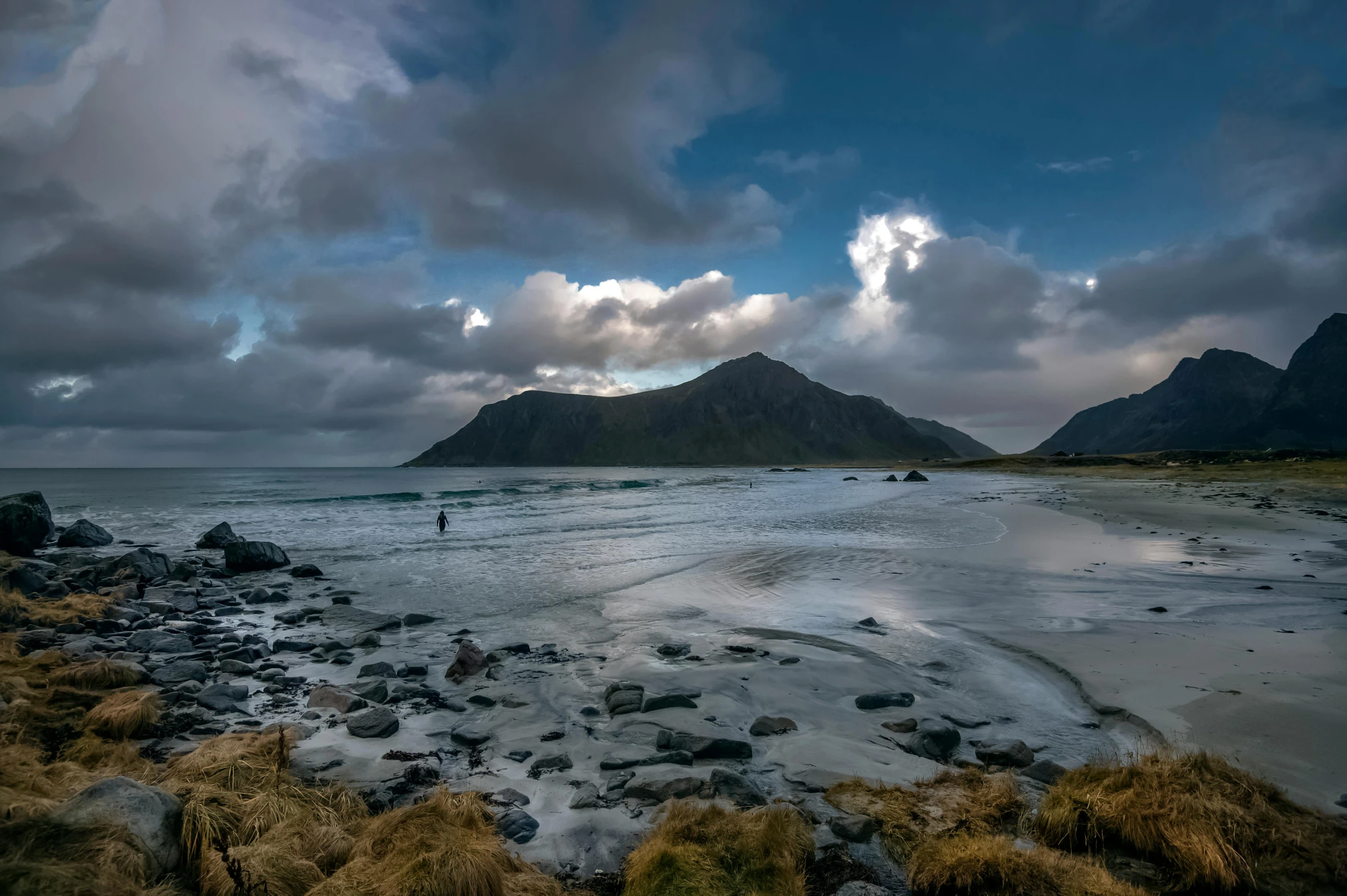 a person standing on a beach next to a body of water, by Harald Giersing, unsplash contest winner, hurufiyya, dramatic mountains in background, under blue clouds, hammershøi, panoramic