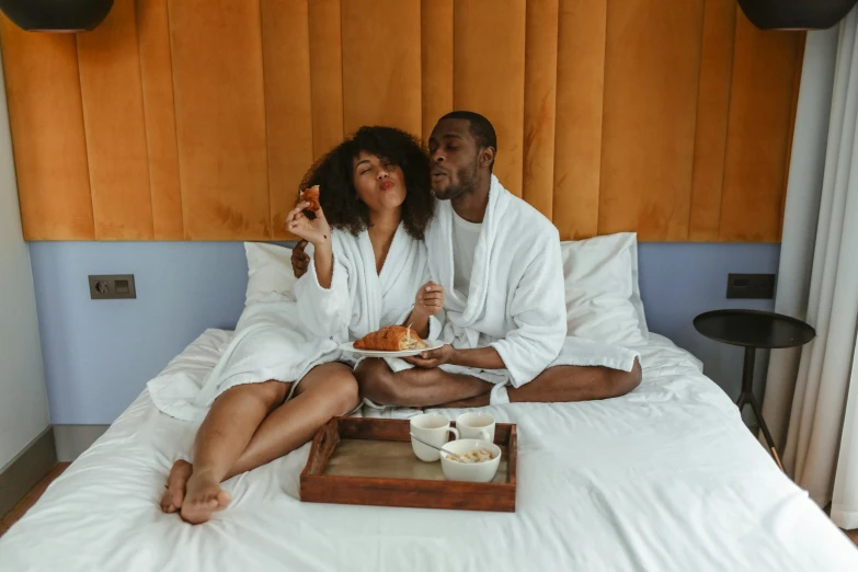 a couple of people that are sitting on a bed, pexels contest winner, yummy, wearing robe, dark-skinned, having a snack