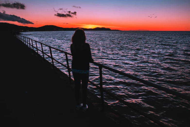 a woman standing on a pier watching the sunset, pexels contest winner, skye meaker, looking out over the sea, it's night time, profile image