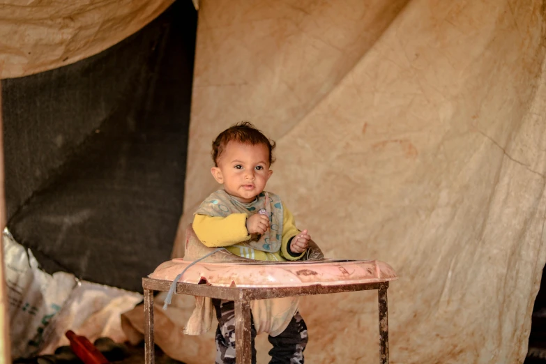 a little boy that is sitting at a table, by Peter Churcher, hurufiyya, temporary emergency shelter, cloth and metal, high quality upload, brown