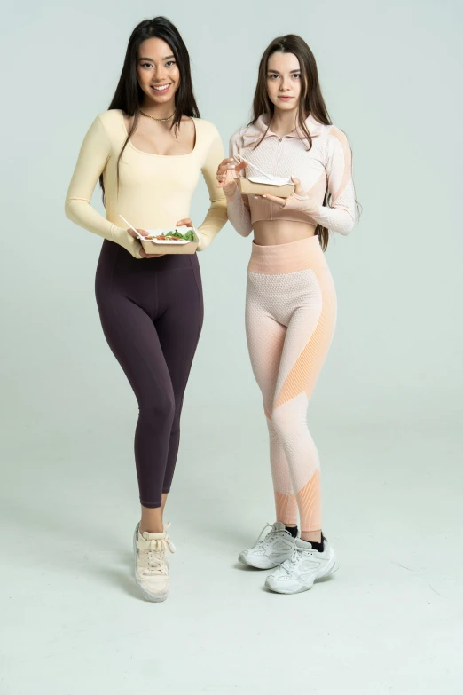 two women standing next to each other holding food, a colorized photo, trending on pexels, leggings, beige, tight outfit, lunging at camera :4