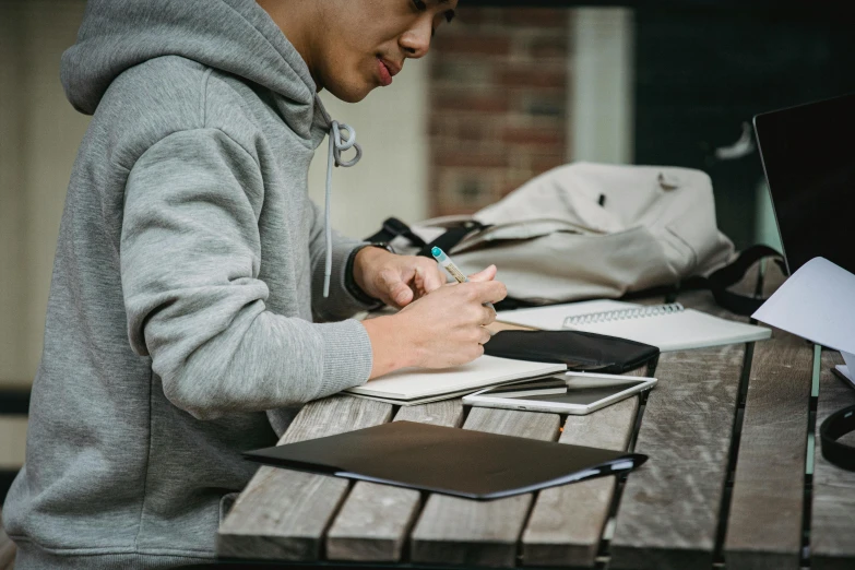 a man sitting at a table writing on a piece of paper, pexels contest winner, academic art, wearing a grey hooded sweatshirt, holding notebook, lachlan bailey, bao pham