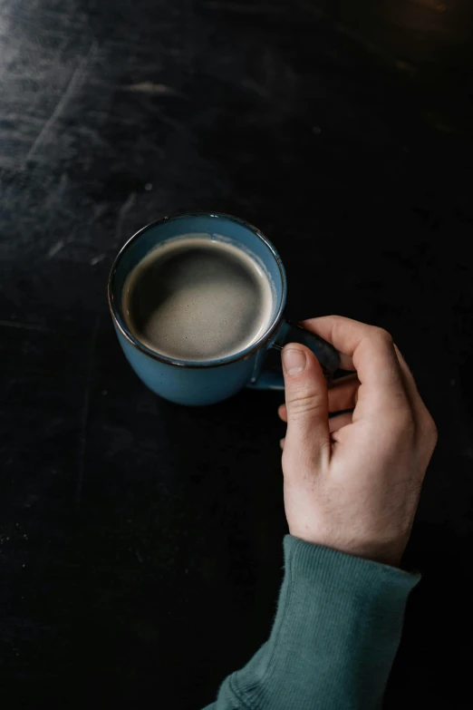 a person holding a cup of coffee on a table, blue and grey, against a deep black background, jen atkin, multiple stories